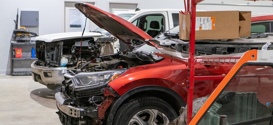 Two severely damaged vehicles in a professional garage. A red SUV has its hood open; a white truck is behind it.