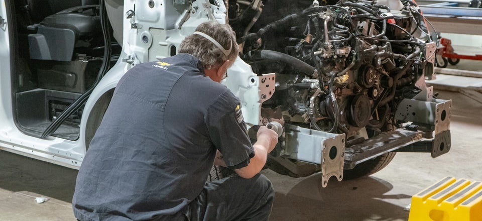 Mechanic with a respirator sanding the front frame of a disassembled white Chevrolet van.